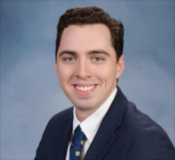 Headshot of Thomas wearing a navy blue blazer, white shirt, and patterned tie.